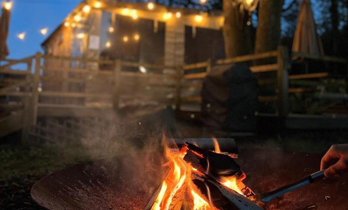 Its night time and a small fire is burning in a fire bowl with a hand stoking the logs. In the background is a wooden cabin lit up by string lights